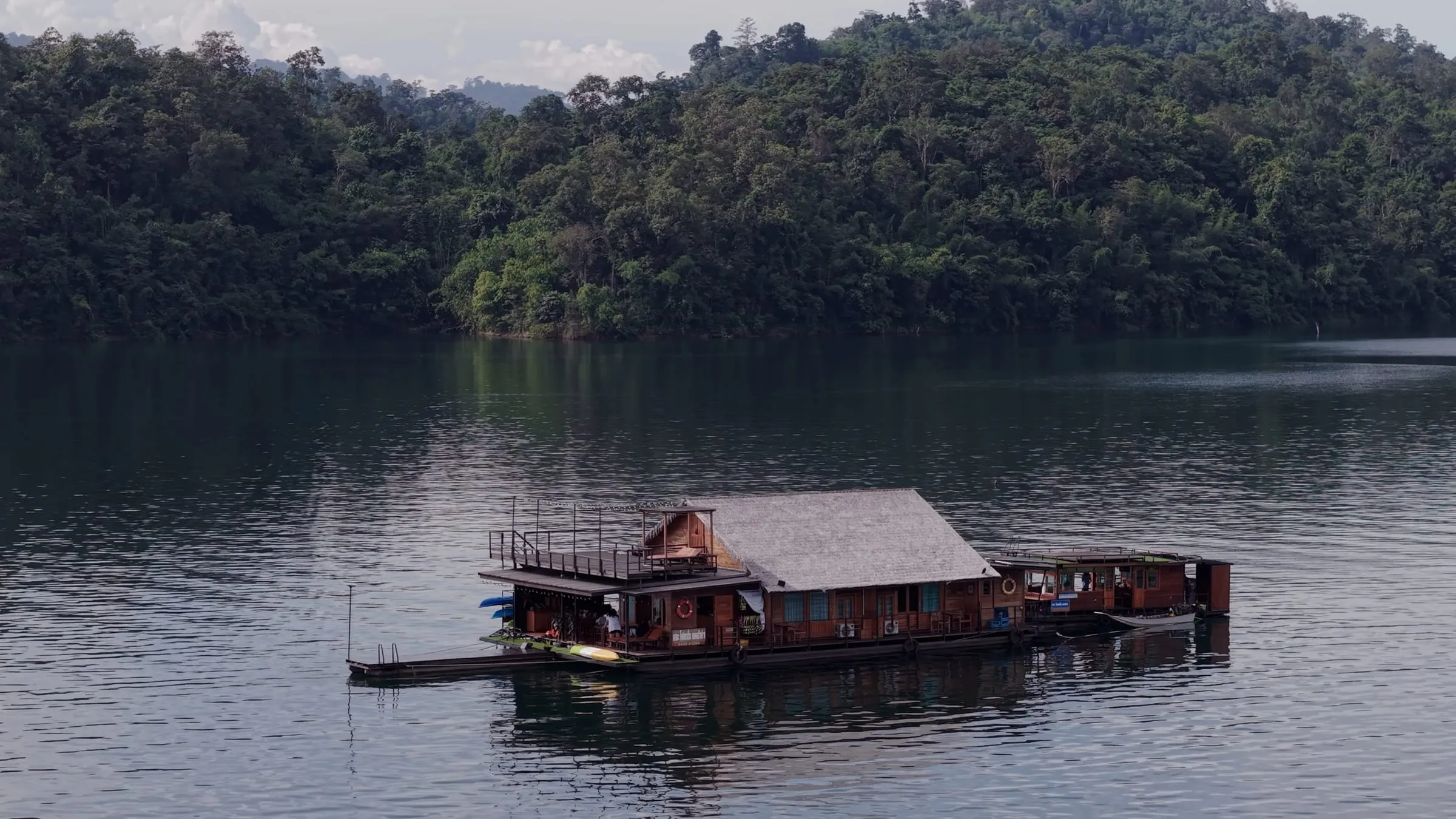 Aerial view of a wooden floating house moored on Khao Laem Lake, ringed by forested mountains.