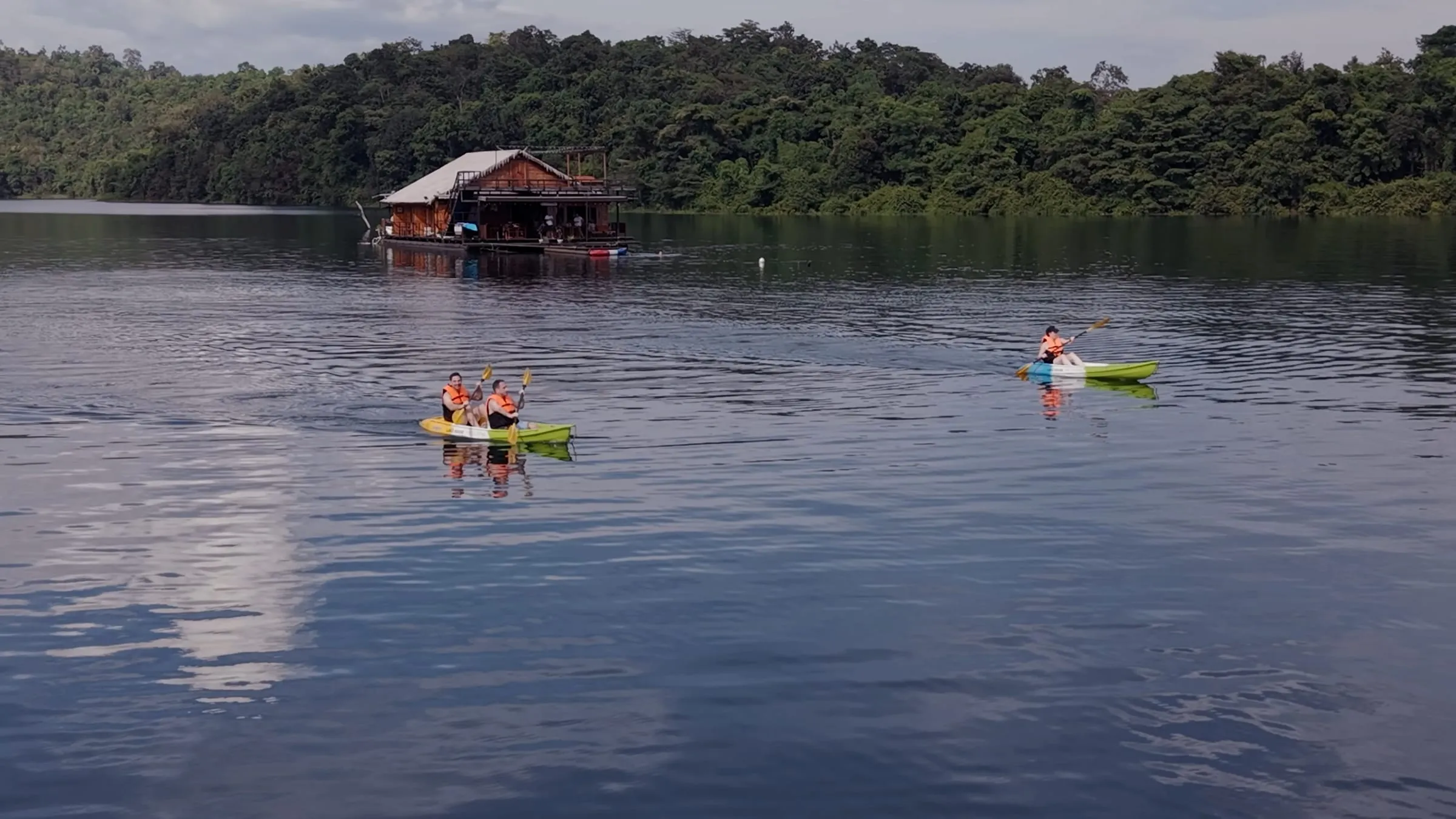 A wooden floating house with two kayaks moored on the calm waters of Khao Laem Lake.