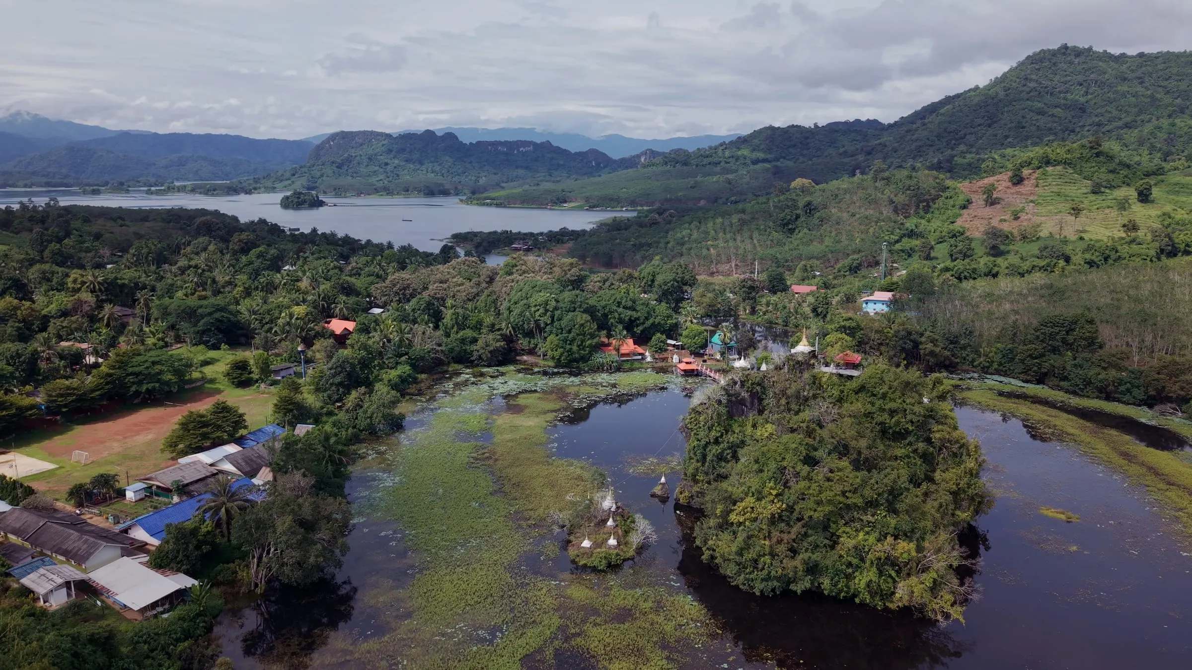 Aerial view of floating houses on a misty arm of Khao Laem Lake among forested hills.